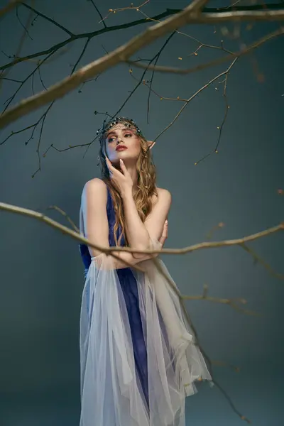 Eine junge Frau in einem blau-weißen Kleid steht anmutig neben einem Baum in einer märchenhaften Kulisse. — Stockfoto