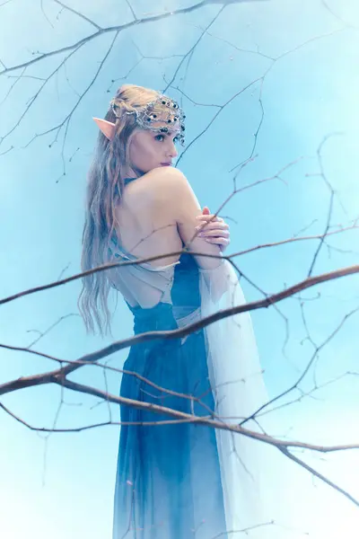 Una joven vestida de azul, parecida a una princesa elfa, se levanta con gracia frente a un majestuoso árbol. - foto de stock