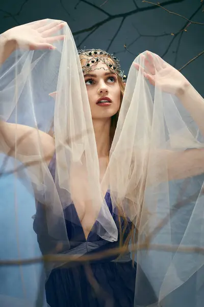 Una mujer joven encarna un cuento de hadas mientras se encuentra en un estudio con un impresionante vestido azul con un velo sobre su cabeza. - foto de stock