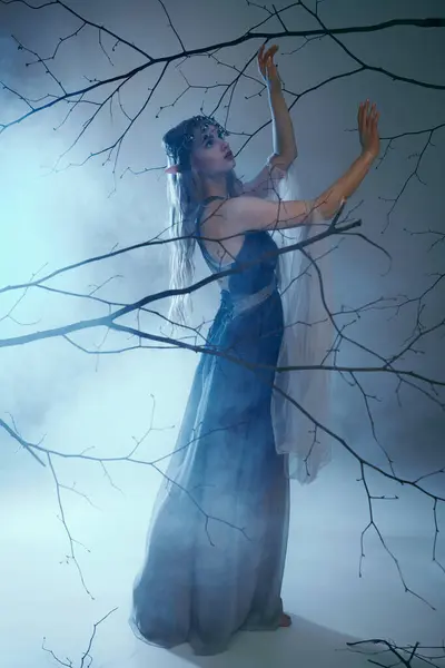 Молодая женщина в синем платье грациозно стоит перед величественным деревом, олицетворяя сущность эльфийской принцессы. — стоковое фото