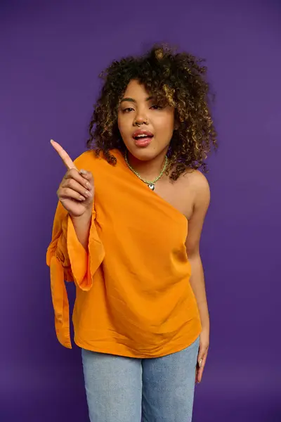 Beautiful African American woman in an orange top pointing at the camera against a vibrant backdrop. — Stock Photo