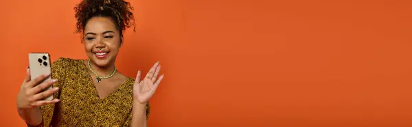 Красивая афроамериканка в стильной одежде держит мобильный телефон перед оранжевой стеной. — стоковое фото
