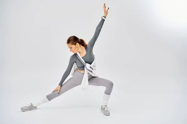 Спортивная молодая женщина в сером и белом наряде изящно практикует позу йоги на сером фоне. — стоковое фото