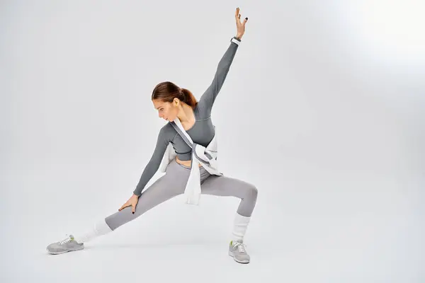 A sporty young woman in grey and white attire gracefully practices a yoga pose on a grey background. — Stock Photo