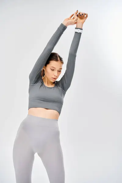 Спортивная молодая женщина в серой спортивной одежде наносит удар в позу йоги с силой и равновесием на сером фоне. — стоковое фото