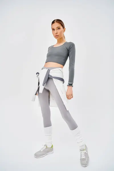 Une jeune femme sportive en tenue grise et blanche prenant une pose confiante sur fond gris. — Photo de stock