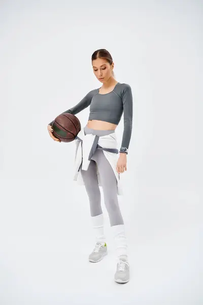 Спортивная молодая женщина изящно держит баскетбольный мяч в правой руке, одетая в активную одежду на сером фоне. — стоковое фото