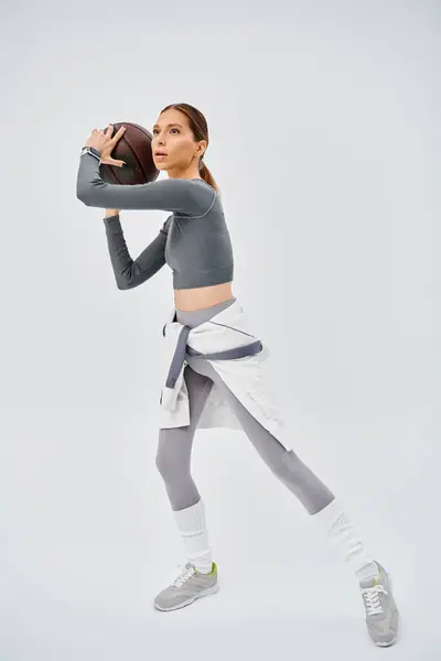Active young woman in sportswear confidently holds a basketball in her right hand against a grey background. — Stockfoto