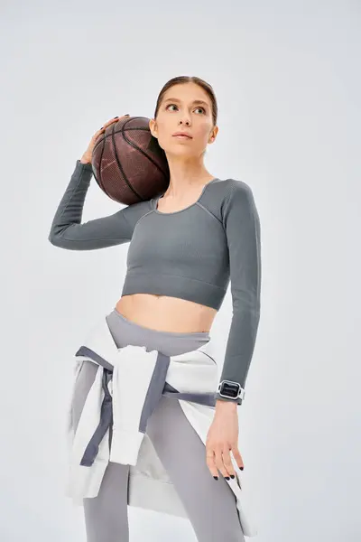 Спортивна молода жінка впевнено тримає баскетбол в правій руці, демонструючи свою спортивну майстерність на сірому фоні. — стокове фото