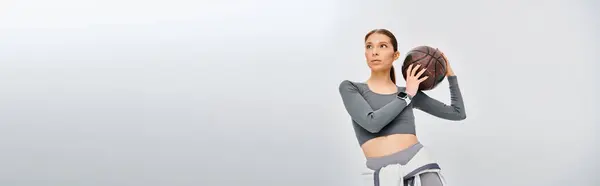 A sporty young woman elegantly holds a basketball in her right hand against a grey background. — Stock Photo