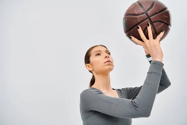 A sporty young woman in active wear holding a basketball up in the air against a grey background. — Stock Photo