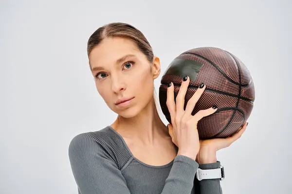Спортивна молода жінка в активному одязі тримає баскетбол над її обличчям на сірому фоні. — стокове фото