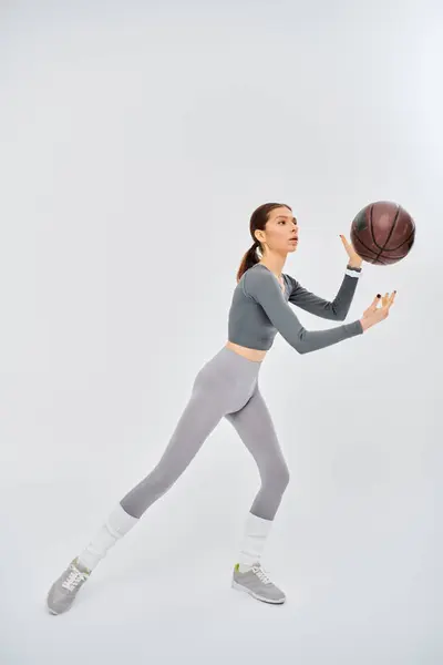 A sporty young woman confidently holds a basketball in her right hand, dressed in active wear against a grey background. — Stockfoto