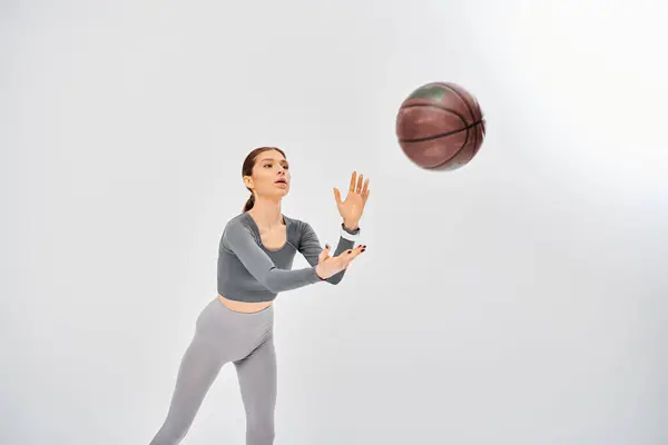 Active young woman in gray top skillfully dribbling basketball in a playful manner on a grey background. — Stockfoto