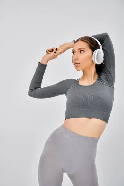 Eine sportliche junge Frau in aktiver Kleidung posiert, während sie über Kopfhörer auf grauem Hintergrund Musik hört.. — Stockfoto