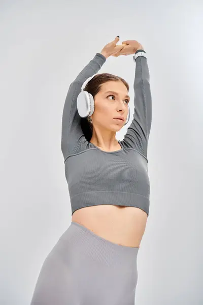 Une jeune femme sportive en tenue active écoutant de la musique à travers des écouteurs, portant un haut gris sur un fond gris. — Photo de stock