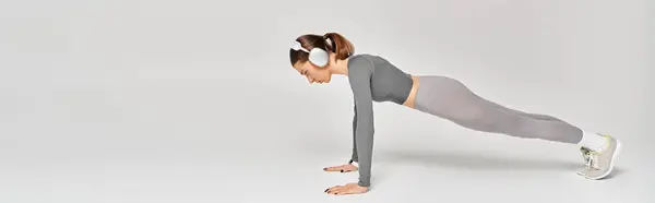 A sporty young woman in active wear demonstrating strength by performing a one-legged push up on a grey background. — Stockfoto