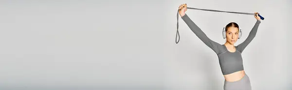 Uma jovem esportiva de camisa cinza segura com confiança uma corda pulando acima de sua cabeça contra um fundo cinza. — Fotografia de Stock