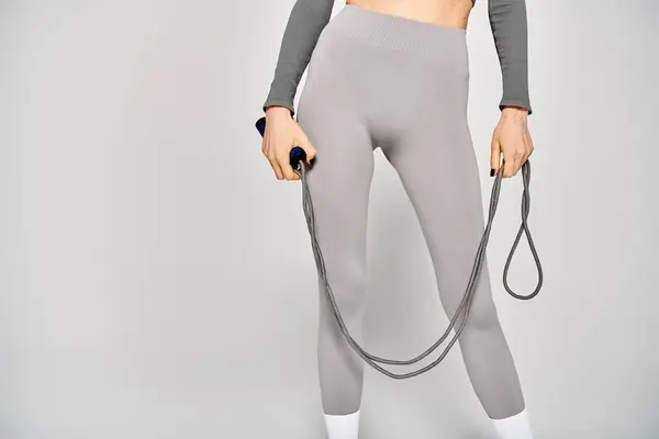 A sporty young woman in grey pants stands confidently, holding a skipping rope against a grey background. — Stockfoto