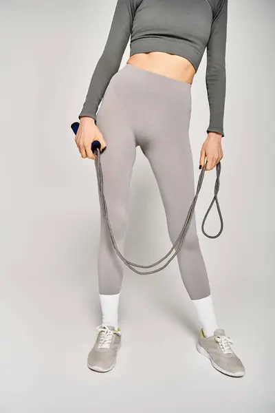 Eine sportliche junge Frau in aktiver Kleidung hält vor grauem Hintergrund ein Springseil in der Hand. — Stockfoto