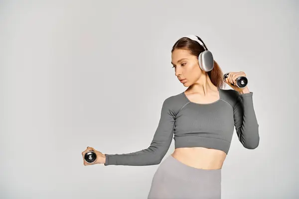 A sporty young woman holds dumbbells while wearing headphones, exuding focus and determination against a grey background. — Stockfoto