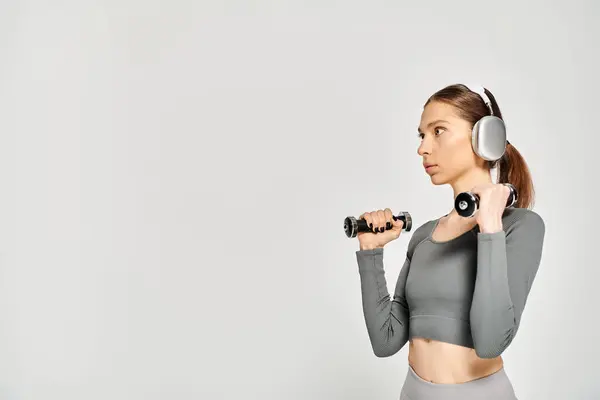 A sporty young woman holding a pair of dumbbells in active wear, showcasing strength and fitness on a grey background. — Stock Photo