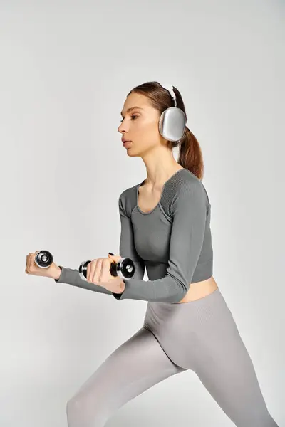 A sporty young woman in active wear is holding a dumbbell while listening to music through headphones on a grey background. — Stock Photo