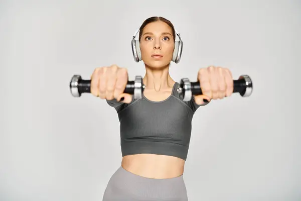 A sporty young woman in active wear, wearing headphones, holds a dumbbell on a grey background. — Stock Photo