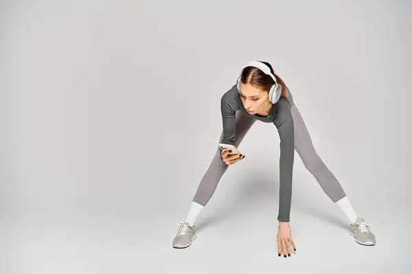 Una giovane donna sportiva in un abito grigio e bianco è focalizzata sul suo schermo del telefono cellulare su uno sfondo grigio. — Foto stock