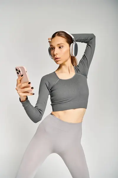 A sporty young woman in active wear, holding a cell phone and wearing headphones, engaged in a phone call on a grey background. — Stockfoto