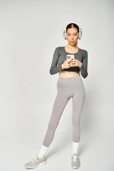 Eine sportliche junge Frau in aktiver Kleidung mit Kopfhörern blickt vor grauem Hintergrund auf ihr Handy.. — Stockfoto