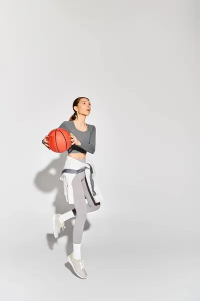 Спортивна молода жінка в активному одязі витончено тримає баскетбол на сірому фоні. — стокове фото