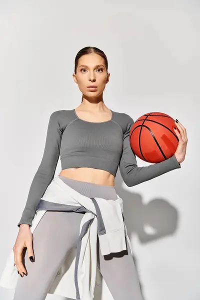 Una joven deportista en ropa deportiva sostiene con elegancia una pelota de baloncesto en su mano sobre un fondo gris. - foto de stock