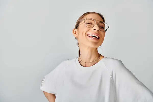 Une jeune femme aux lunettes rayonne brillamment dans une chemise blanche croustillante sur fond gris neutre. — Photo de stock