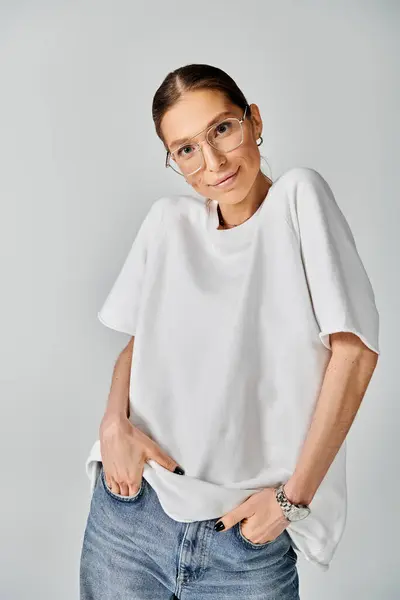 Молодая женщина в белой футболке и очках позирует на сером фоне, излучая элегантность и уверенность. — стоковое фото