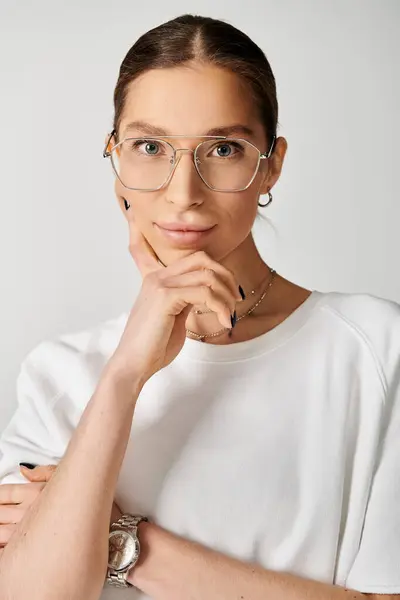 Una joven vestida con una camiseta blanca alcanza una pose, mostrando sus elegantes gafas sobre un elegante fondo gris. - foto de stock