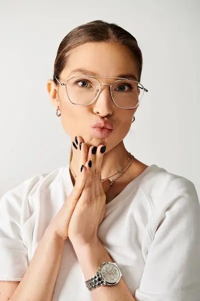 Una mujer joven con una camiseta blanca y gafas está haciendo una cara divertida sobre un fondo gris. - foto de stock