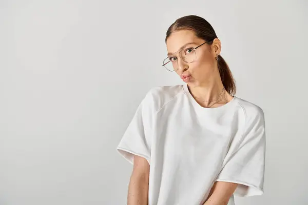 Una joven con gafas y una camisa blanca se levanta sobre un fondo gris. - foto de stock