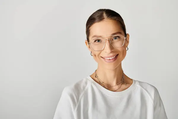 Une jeune femme en t-shirt blanc portant des lunettes sourit directement à la caméra sur un fond gris. — Photo de stock