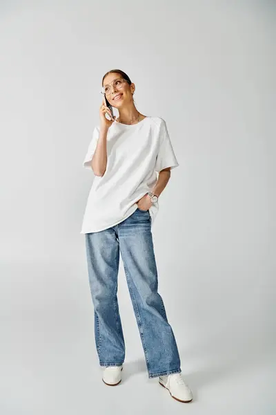 Una joven con camisa blanca y jeans hablando por celular sobre un fondo gris. - foto de stock