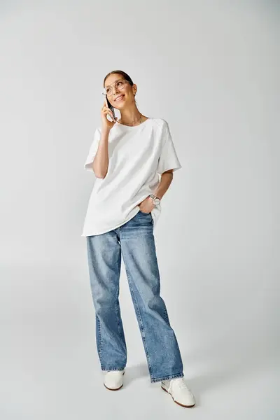 A young woman in a white shirt and jeans talking on a cell phone against a grey background. — Stock Photo