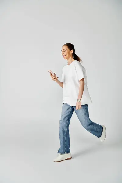 Una joven con una camiseta blanca y gafas camina mientras se enfoca en la pantalla de su teléfono celular. - foto de stock