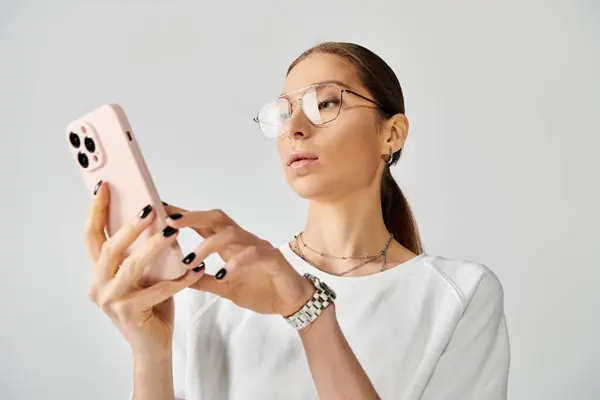 Una joven con estilo en gafas que sostiene un teléfono celular sobre un fondo gris. - foto de stock