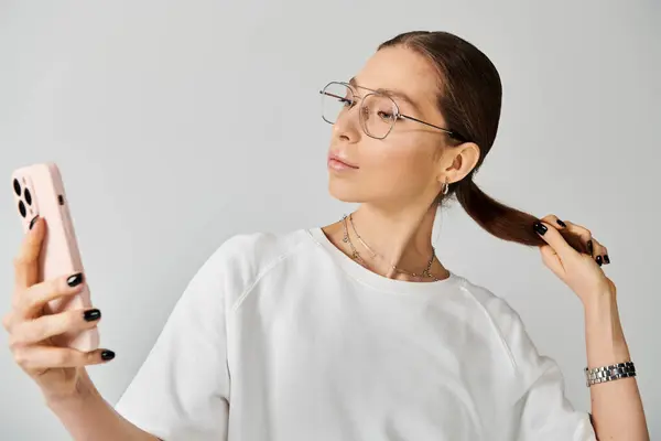 Una joven con una camiseta blanca y gafas sostiene un teléfono celular, sus ojos enfocados en la pantalla. — Stock Photo
