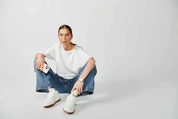 Una giovane donna con una t-shirt bianca e occhiali è seduta a terra mentre tiene in mano uno smartphone — Foto stock