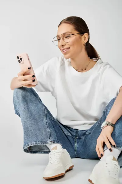 Une jeune femme portant un t-shirt blanc et des lunettes s'assoit sur le sol, tenant paisiblement un téléphone portable. — Photo de stock