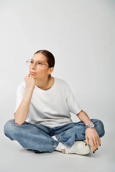 Uma jovem mulher em uma camiseta branca e óculos senta-se no chão, queixo descansando na mão, perdido em pensamento. — Fotografia de Stock