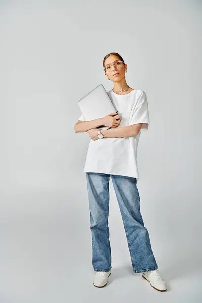 Una joven mujer está de pie con confianza con el ordenador portátil, con una camiseta blanca y gafas sobre un fondo gris. - foto de stock