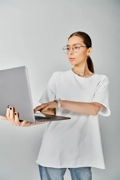 Una joven mujer sostiene con confianza un portátil en la mano, con una camiseta blanca y gafas sobre un fondo gris. - foto de stock