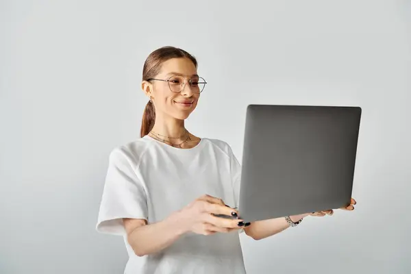 Una joven con una camiseta blanca y gafas sosteniendo una computadora portátil en sus manos sobre un fondo gris. - foto de stock